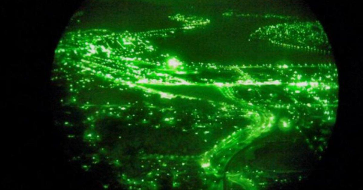 Aerial view of a city at night seen through night vision goggles, showing green-tinted terrain with illuminated roads and buildings